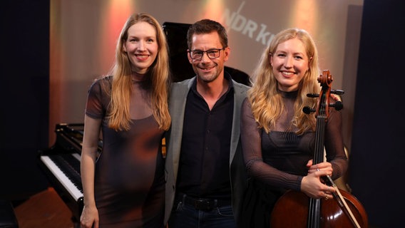 Ein Mann steht inmitten zweier junger Frauen, eine von ihnen hält ein Cello in der Hand. © Ekaterina Shurygina / NDR Foto: Ekaterina Shurygina / NDR