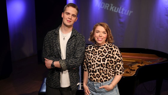 Ein großer, junger Mann mit einem weißen T-Shirt und einem grau, schwarz, gemusterten Hemd steht neben einer Frau mit einem Leoprint-Shirt. Beide lachen in die Kamera. © Ekaterina Shurygina / NDR Foto: Ekaterina Shurygina / NDR