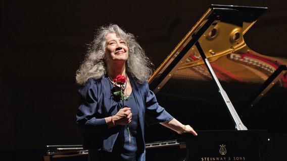 Eine Frau mit langen grauen Haaren (die Pianistin Martha Argerich) steht mit einer Rose in der Hand an einem Konzertflügel. © Daniel Dittus Foto: Daniel Dittus