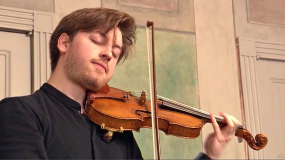 Ein junger Mann mit schwarzem Hemd spielt im Stehen mit geschlossenen Augen eine kostbare Geige © NDR Screenshot Foto: Inessa Baur