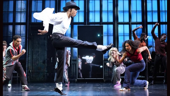 Ein Mann mit Hut tanzt mit erhobenem Bein zwischen Tänzerinnen und Tänzern - Szene aus dem Michael-Jackson-Musical "MJ". © picture alliance / dpa Foto: Marcus Brandt