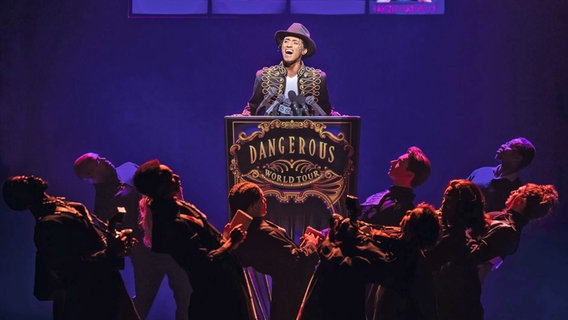 Ein Mann mit Hut steht an einem Pult, darauf steht "Dangerous", davor stehen Menschen - Szene des Musicals "MJ" in Hamburg © Stage Entertainment /Matthew Murphy Foto: Matthew Murphy