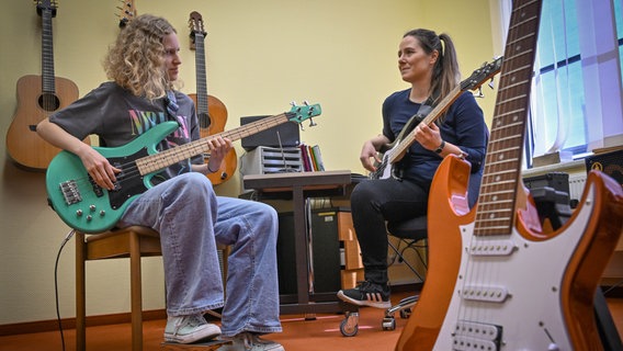 Zwei Frauen sitzen sich gegenüber und spielen E-Gitarre. An der Wand hängen mehrere Akustikgitarren. © picture alliance/dpa | Patrick Pleul 