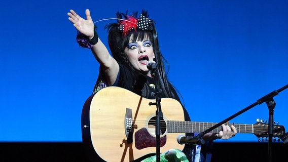 Sängerin Nina Hagen sitzt auf einem Hocker vor einem blauen Hintergrund mit einer Gitarre auf dem Schoß. Ihre rechte Hand hat sie jugelnd nach oben gerissen. © picture alliance / Jens Kalaene/dpa-Zentralbild/dpa Foto: Jens Kalaene