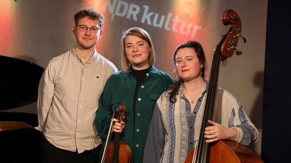 Zwei junge Frauen, die eine hält eine Geige und die andere einen Kontrabass in der Hand, stehen neben einem großen Mann. © Ekaterina Shurygina / NDR Foto: Ekaterina Shurygina / NDR