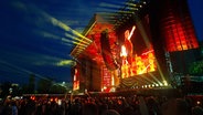 Eine rötlich beleuchtete Bühne mit einer großen Leinwand - darauf zu sehen ist der Sänger Brian Johnson. Ein Publikum jubelt ihm zu. © NDR Foto: Uli Kniep