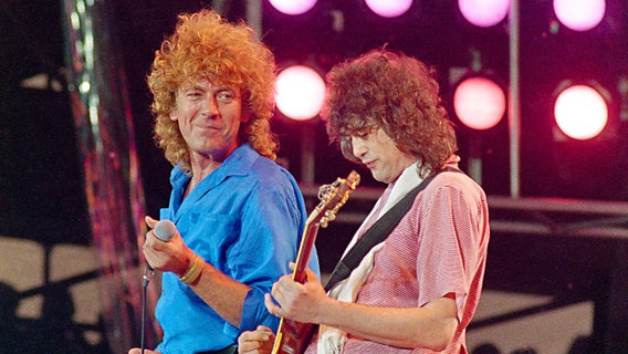 Led Zeppelin-Sänger Robert Plant (links) und -Gitarrist Jimmy Page bei einem Auftritt auf einem Live Aid Konzert in Philadelphia im Juli 1985 auf der Bühne. Page spielt Gitarre, während ihn Sänger Plant anschaut und lächelt. © picture alliance / AP Photo | AMY SANCETTA Foto: picture alliance / AP Photo | AMY SANCETTA