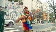 Ein Mann mit einem bunten T-Shirt hält eine Ukulele spielbereit und steht auf der Straße in einer Stadt. © dpa Bildfunk 