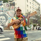 Ein Mann mit einem bunten T-Shirt hält eine Ukulele spielbereit und steht auf der Straße in einer Stadt. © dpa Bildfunk 