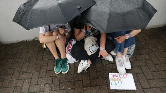 Drei junge Fans sitzen beim Konzert der US-Sängerin Taylor Swift im Volksparkstadion unter ihren Regenschirmen vor dem Stadion. © Christian Charisius/dpa Foto: Christian Charisius