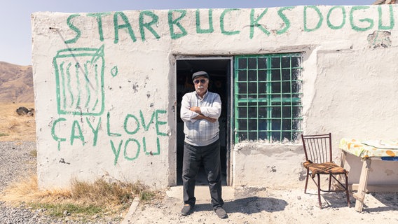 Ein älterer Mann steht mit verschränkten Armen für einem Lehmhaus mit der Aufschrift "Starbucks" © NDR.de/Christina Grob Foto: Christina Grob