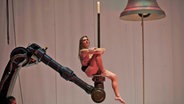 Eine kaum verhüllte Darstellerin sitzt auf einem Kran, vor ihr hängt eine riesige Glocke, über ihr ist eine angezündete Kerze (Szene aus der Oper "Sancta" in Stuttgart) © Matthias Baus/Staatsoper Stuttgart/dpa Foto: Matthias Baus