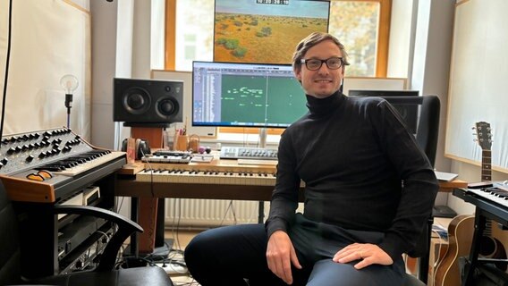 Der Filmmusik-Komponist Sascha Blank sitzt im Rollkragenpullover auf einem Stuhl in seinem Studio und lächelt in die Kamera. Im Hintergrund sind diverse Musik- und Elektroinstrumente sowie Bildschirme zu sehen, auf denen Musikbearbeitungsprogramme und Videos geöffnet sind. © NDR Foto: Martin Möller