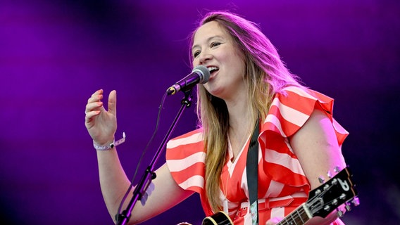 Eine Frau steht mit langen Haaren und einem rot-weiß-gestreiften Kleid auf der Bühne und singt in ein Mikrofon. Sie hat eine Gitarre um den Hals. © picture alliance/dpa | Britta Pedersen Foto: Britta Pedersen