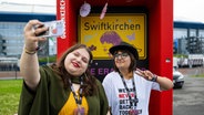 Zwei junge Frauen machen einen Selfie vor dem Plakate "Swiftkirchen" vor der Arena in Gelsenkirchen, wo Taylor Swift auftreten wird ©  Guido Kirchner/dpa +++ dpa-Bildfunk +++ Foto: Guido Kirchner