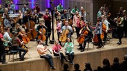 Ein bunt gekleidetes Orchester sitzt und steht spielend auf einer Bühne. © Bernd Possardt 