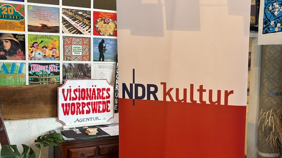 Links hängen Plattencover an der Wand. Darunter steht ein Schild mit der Aufschrift "Visionäres Worpswede". Rechts steht ein NDR Kultur Aufsteller. © NDR Kultur Foto: Tom Holste, Cornelius Kob