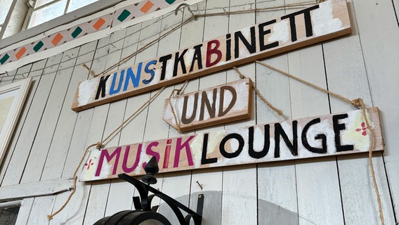 An der Holzwand hängen drei Holzschilder untereinander. Darauf stehen: Kunstkabinett und Musiklounge. © NDR Kultur Foto: Tom Holste, Cornelius Kob