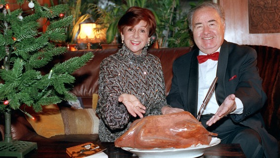 Der Schauspieler und Conferencier Heinz Quermann mit Margot Ebert. Die Beiden zeigen auf eine verpackte Gans die auf dem Tisch steht. Links von Ihnen steht ein Weihnachtsbaum. © picture-alliance / dpa | Zentralbild Foto: -