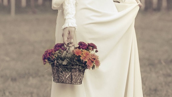 Eine Frau in einem viktorianischem Kleid läuft über eine Wiese mit einem Korb Blumen in der Hand. © imago Foto: Joana Kruse