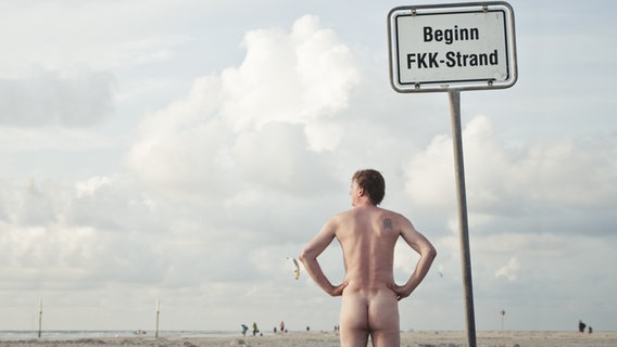 Ein nackter Mann schaut in die Ferne, neben ihm ein Schild mit der Aufschrift "Beginn FKK-Strand". © una.knipsolina / photocase.de Foto: una.knipsolina