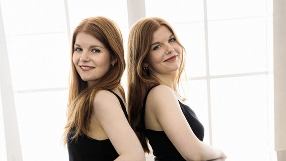 Zwei Frauen mit roten langen Haaren (Zwillingsschwestern) stehen Rücken an Rücken und schauen freundlich nach vorne. © Clara & Marie Becker 