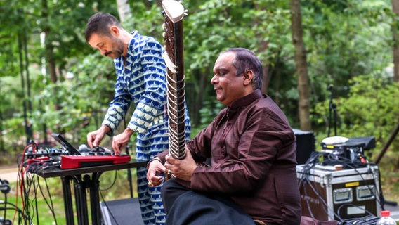 Zwei Musiker bei einem Open Air Auftritt. Einer spielt Sitar, der andere steht im Hintergrund an einem Mischpult. © imago images / Votos-Roland Owsnitzki Foto: Votos-Roland Owsnitzki