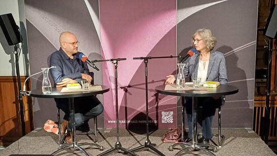 Ein Mann mit Glatze und Brille und eine Frau mit kinnlangen grauen Haaren sitzen auf einer Bühne an Tischen. © NDR Foto: Alexander Solloch