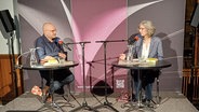 Ein Mann mit Glatze und Brille und eine Frau mit kinnlangen grauen Haaren sitzen auf einer Bühne an Tischen. © NDR Foto: Alexander Solloch