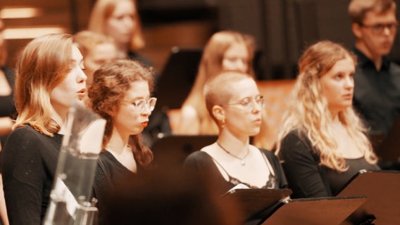 Vier junge Frauen in schwarzer Kleidung sehen auf Notenblätter und singen. © NDR 