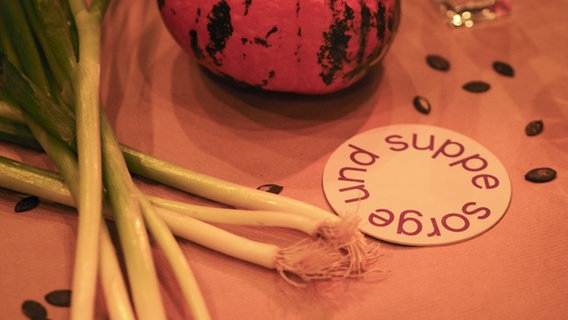 Bierdeckel mit der Aufschrift "Sorge und Suppe" liegen auf einem Tisch, daneben sind Lauchzwiebel, Kürbis und eine Kerze. © NDR/Margarita Ilieva Foto: Margarita Ilieva