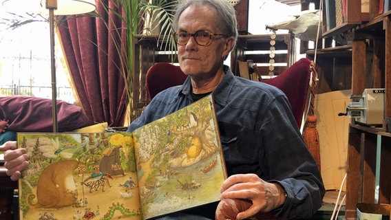 Sven Nordqvist sitzt in einem roten Sessel und zeigt ein aufgeklapptes Kinderbuch. © NDR 