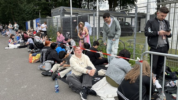 Taylor Swift Fans stehen und sitzen teilweise mit Glitzeroutfit, teilweise ohne in der Schlange vor dem Stadion. © Julia Hercka / NDR Foto: Julia Hercka / NDR