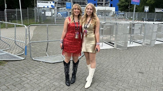 Zwei junge Frauen stehen vor dem Volksparkstadion und tragen kurze Glitzerkleider und hohe Stiefel. © Julia Hercka / NDR Foto: Julia Hercka / NDR