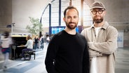 Mark Forster und Igor Levit stehen in einer Bahnhofshalle und lächeln in die Kamera, während eine Person im Hintergrund auf einem Klavier spielt. © Kalimba 