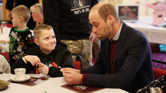 Prinz William sitzt mit einem Jungen am Tisch und verzieht spielerisch das Gesicht. © Richard Pohle/The Times/PA Wire/dpa 