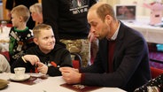 Prinz William sitzt mit einem Jungen am Tisch und verzieht spielerisch das Gesicht. © Richard Pohle/The Times/PA Wire/dpa 