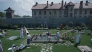 Eine große Familie springt auf der grünen Wiese um einen kleinen Pool herum - dahinter ist ein KZ zu erkennen. Szene aus dem britisch-polnischen Spielfilm "The Zone of Interest" von Jonathan Glazer © Leonine 