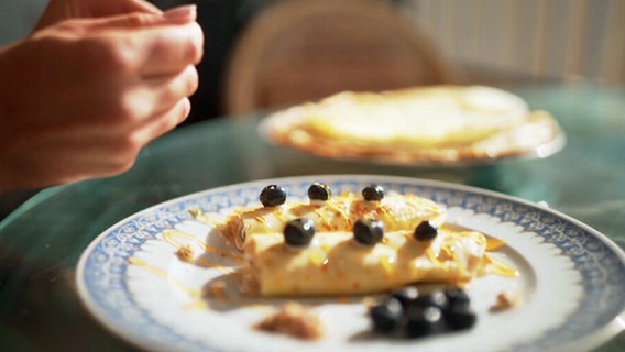 Ein Teller mit gebackenen Blinis, darüber eine weibliche Hand. © NDR 