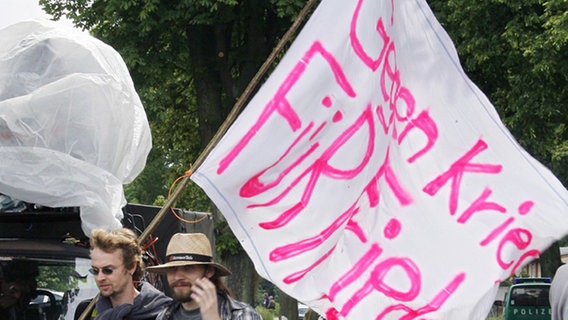 Mit einer Fahne mit der Aufschrift "Gegen Krieg für Frieden" kommen in Weitendorf zwei Demonstranten zu der abgesperrten Zugangsstraße zum Flughafen Rostock-Laage. © dpa 