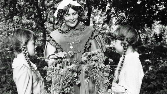 Schulfräulein Haubold (Katherina Schmidt-Lüdge) bemüht sich, mit den beiden Schwestern Lining (Tanja Kracht, links) und Minig (Nicole Behrends) die Sorten der gepflückten Blumen zu bestimmen. © NDR/Hassenstein 