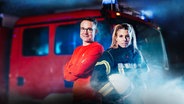 Ein Mann in rotem Pullover und eine Frau in Feuerwehruniform mit Helm stehen vor einem roten Feuerwehrauto mit Blaulicht und Rauch im Hintergrund. © NDR Foto: Andrea Seifert