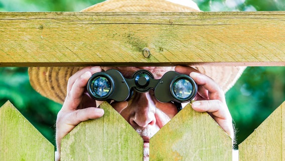 Ein Nachbar schaut mit einem Fernglas neugierig durch einen Zaun. © Rainer Fuhrmann / photocase.de Foto: Rainer Fuhrmann / photocase.de