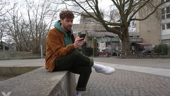 Screenshot aus einer Y-Kollektiv-Reportage: Reporter Frederick Fleig sitzt mit seinem Handy in der Hand auf einer Mauer. © YouTube / Y-Kollektiv Foto: Screenshot