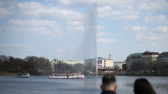 Ein Wasserstrahl der Alsterfontäne schießt auf der Binnenalster in Hamburg in die Luft, im Vordergrund sitzen zwei Personen. Bis zum November wird der 50 Meter hohe Wasserstrahl nun täglich von 9.00 bis 24.00 Uhr sprudeln. © picture alliance/dpa Foto: David Hammersen