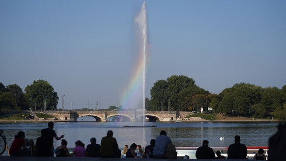 Die Gischt der Alsterfontäne bildet Regenbogenfarben, während am Jungfernstieg Passanten auf die Binnenalster blicken. © picture alliance / dpa Foto: Marcus Brandt