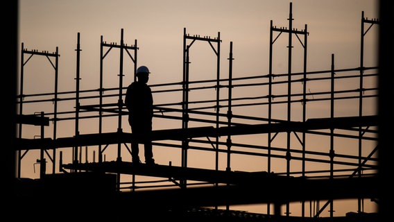 Die Silhouette eines Bauarbeiters zeichnet sich auf einer Baustelle vor dem verfärbten Morgenhimmel ab. © Julian Stratenschulte/dpa 