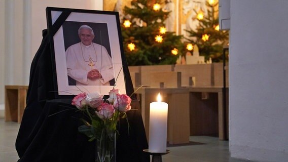 Ein Foto von Papst Benedikt XVI. steht neben Rosen und einer Kerze im Hamburger St. Marien-Dom. © dpa Foto: Marcus Brandt