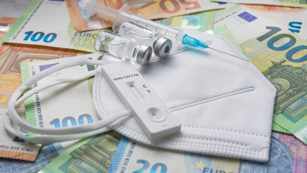 Ein Corona-Schnelltest, eine Maske, zwei Impfstoffflaschen und eine Spritze liegen auf mehreren Euro-Scheinen. (Symbolfoto)