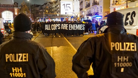 Linksautonome demonstrieren in Hamburg gegen die Polizei. Auf einem Transparent steht "All Cops Are Enemies". Im Vordergrund sind zwei Polizisten zu sehen. © picture alliance / ABBfoto 